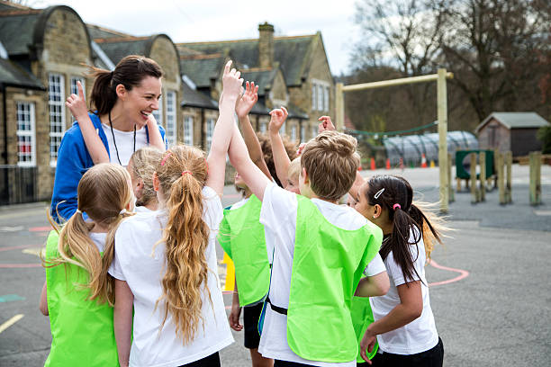 Children in the school playground during a physical education lesson. Some are wearing coloured vests whilst others are just in white t-shirts. They all have their hands up to volunteer for something. The teacher is…